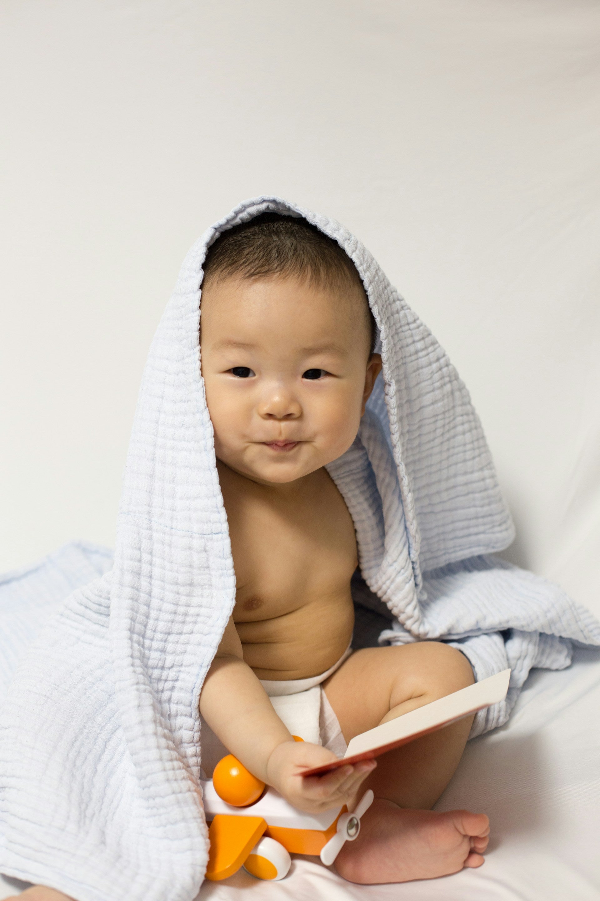 A baby boy with a blanket on his head and a book and wooden toy on his lap.