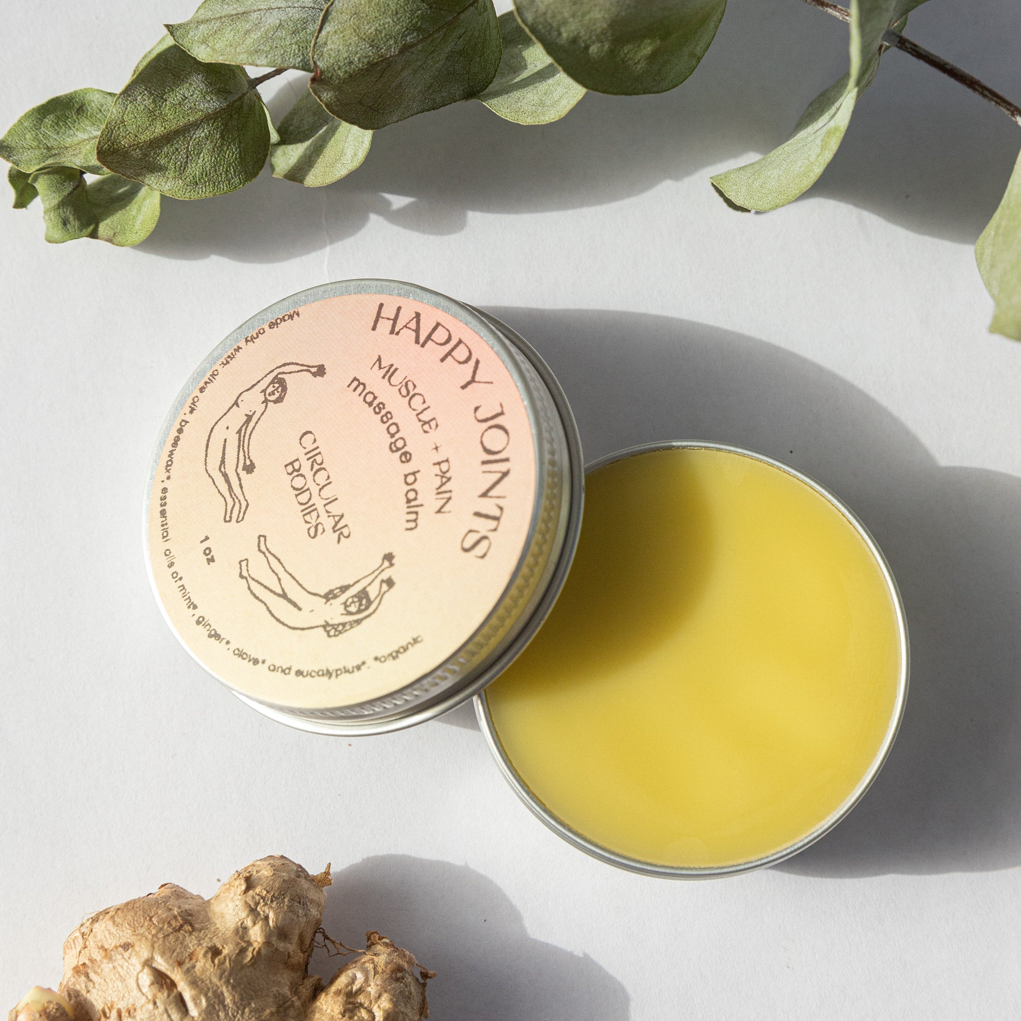 Tin of Happy Joints massage balm with a branch of eucalyptus and ginger root on a light background