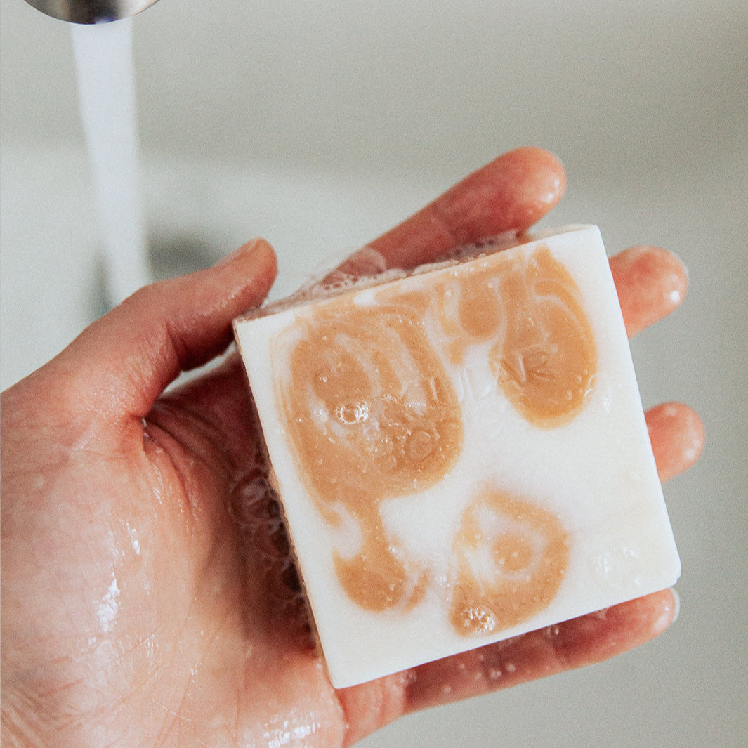 Hand holding a bar of soap with brown specks under running water.